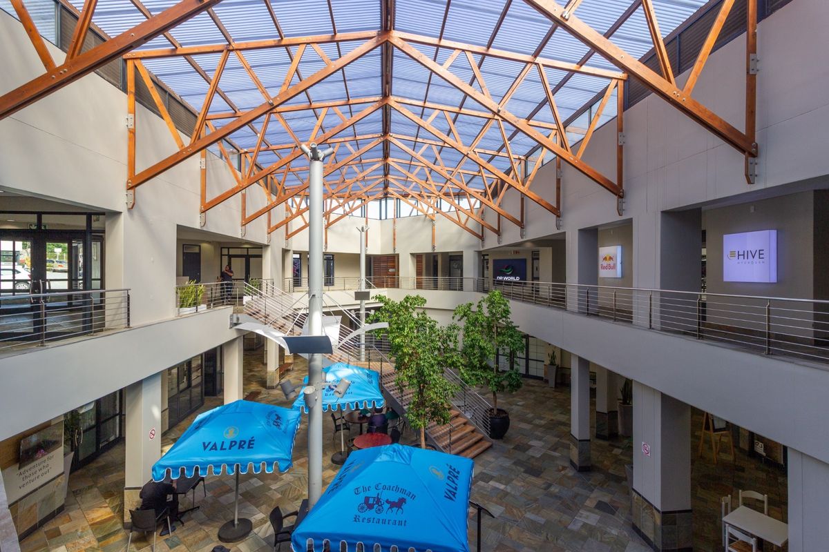 Spacious multi-level retail atrium featuring a high timber truss roof with translucent panels, stone flooring, and mezzanine walkways overlooking a central courtyard area.