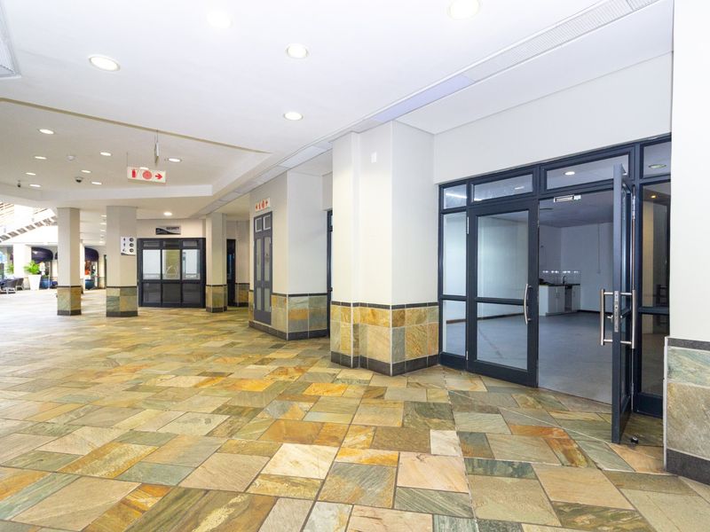 A spacious commercial entry area featuring natural stone tile flooring, recessed lighting, and glass storefront access points within a well-maintained retail complex.