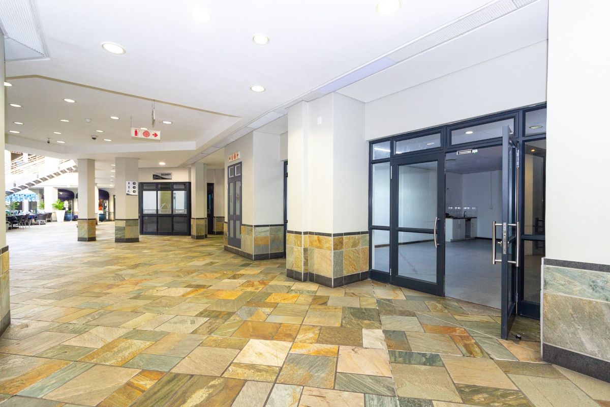 A spacious commercial entry area featuring natural stone tile flooring, recessed lighting, and glass storefront access points within a well-maintained retail complex.