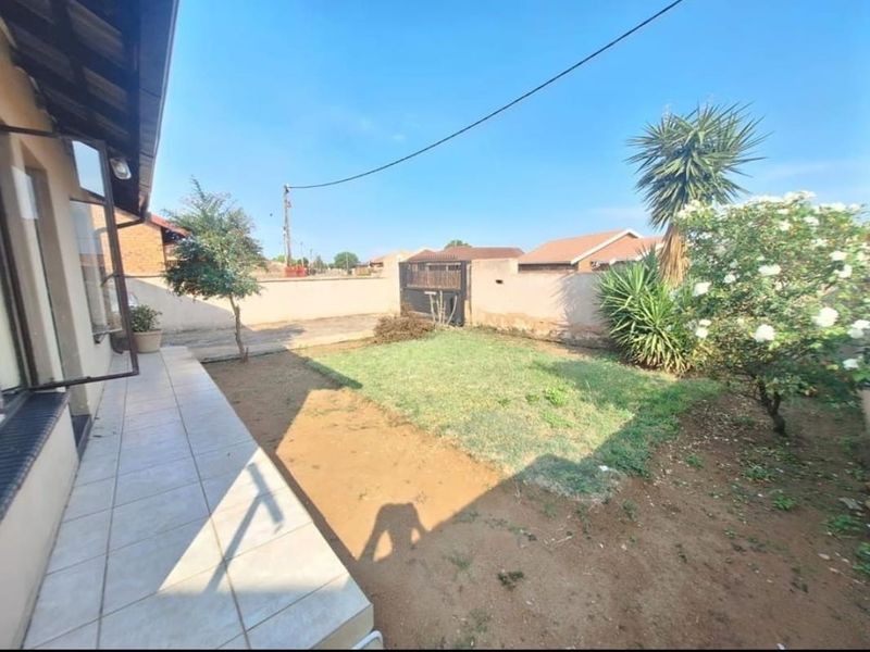 A rear exterior view featuring a tiled patio area, a small lawn, and a perimeter wall with a metal gate.