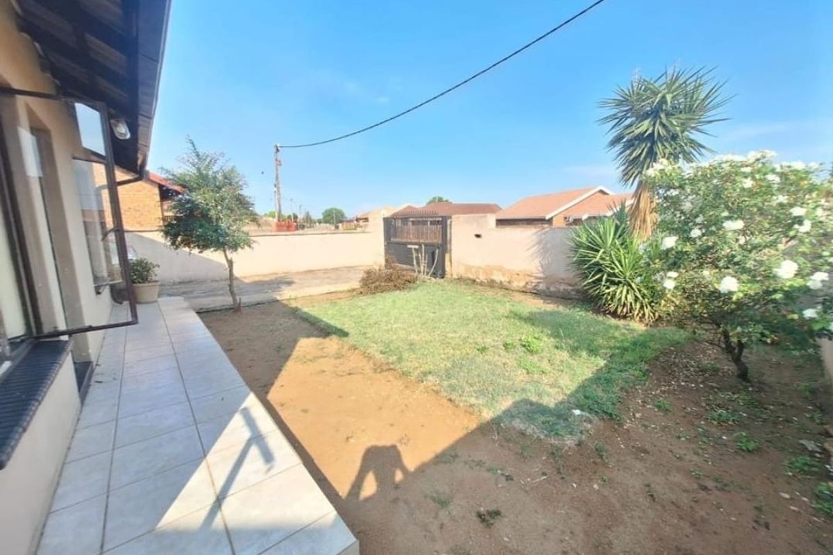 A rear exterior view featuring a tiled patio area, a small lawn, and a perimeter wall with a metal gate.