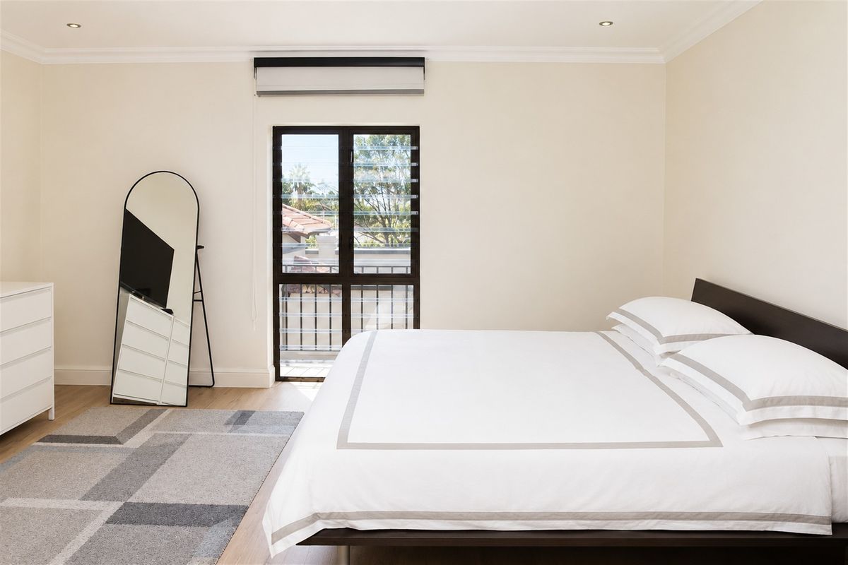 A bright and well-maintained bedroom featuring timber flooring, recessed downlighting, an air conditioning unit, and a louvered window with access to a balcony.