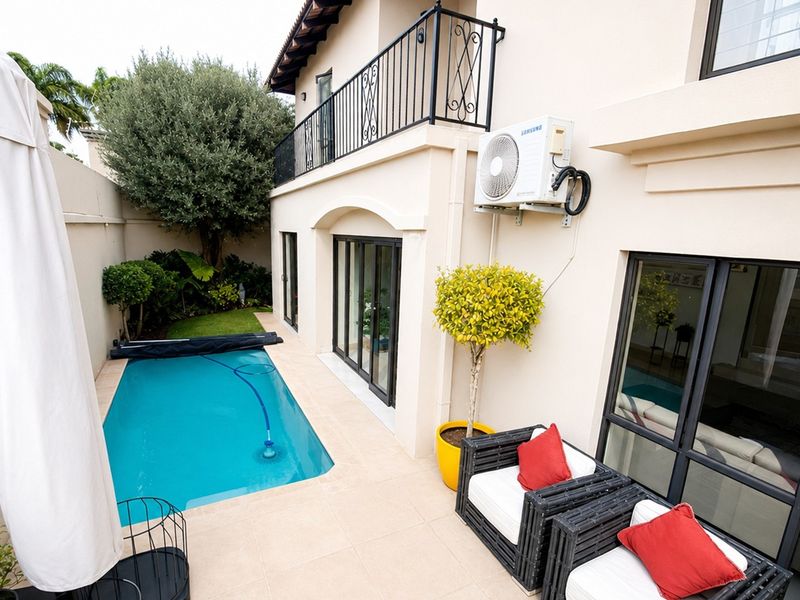 A well-maintained rear patio area featuring a swimming pool, tiled flooring, and a balcony with wrought iron railings overlooking the garden.