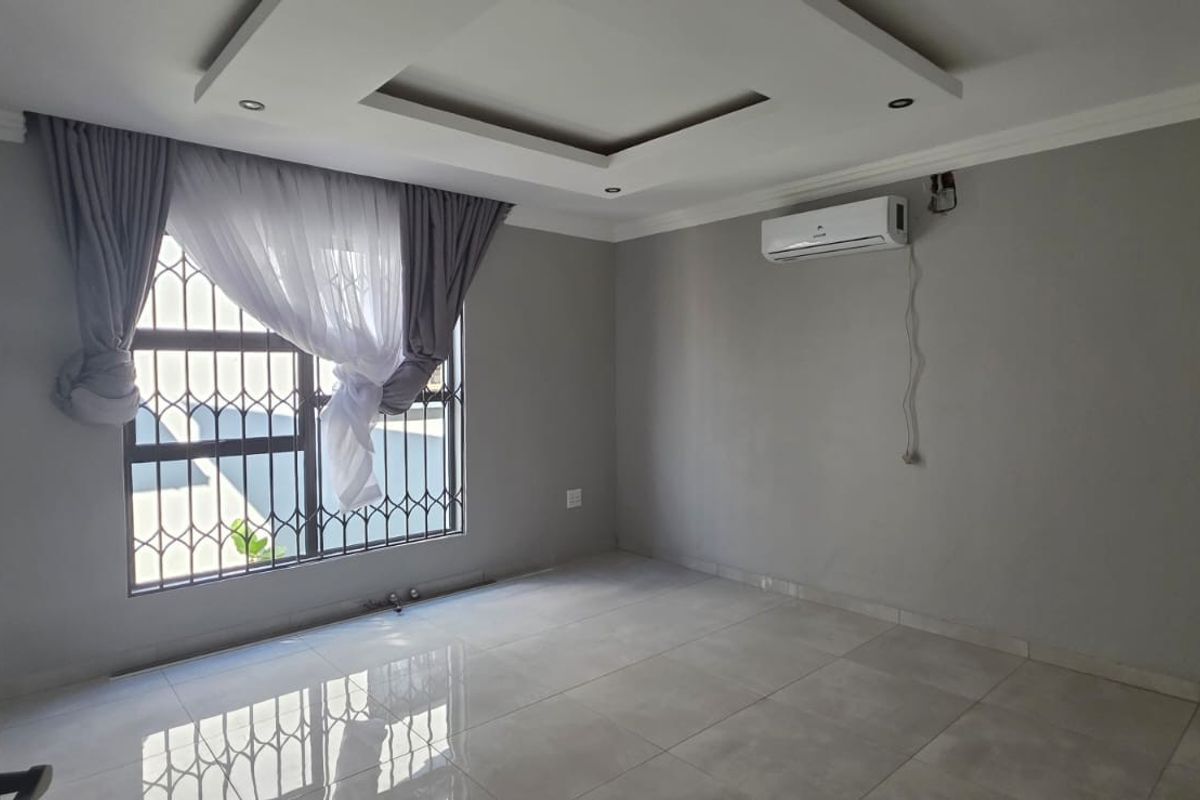 A spacious, empty bedroom featuring tiled flooring, a recessed ceiling design, and a wall-mounted air conditioning unit with security bars on the window.