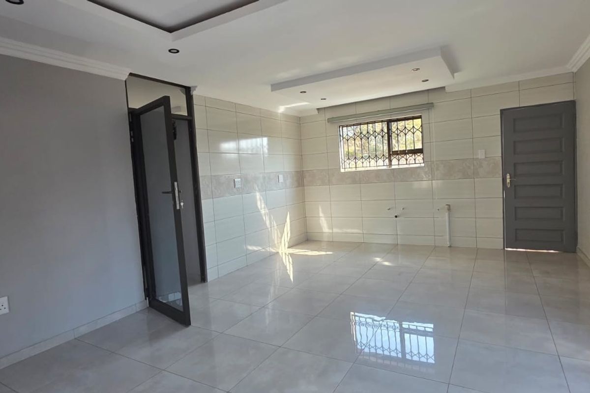 An empty, tiled kitchen area featuring recessed ceiling lighting, security bars on the window, and plumbing connections ready for appliance installation.