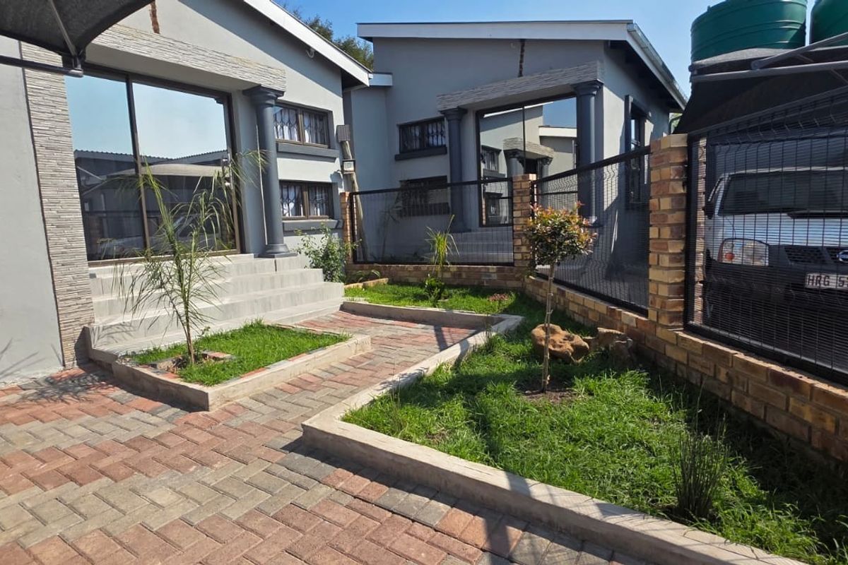 Rear exterior view featuring a paved courtyard, landscaped garden beds, concrete steps leading to sliding glass doors, and secure perimeter fencing.
