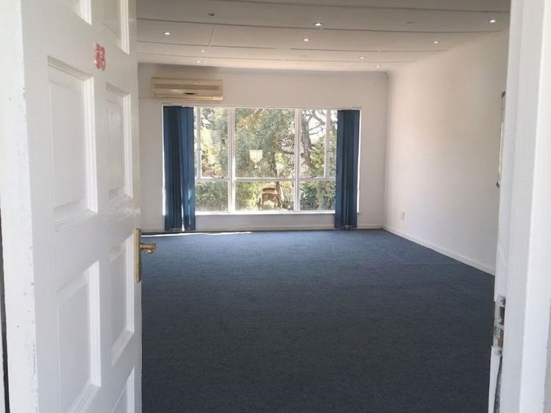 A spacious, empty office area featuring carpeted flooring, recessed downlighting, and a split-system air conditioning unit with large windows providing natural light.