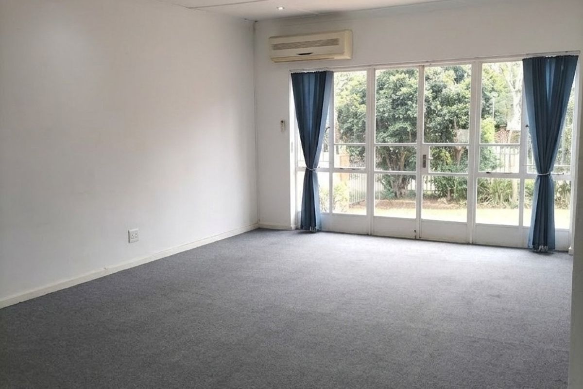 A spacious, empty living area featuring grey carpeting, recessed downlights, a wall-mounted air conditioning unit, and large glass sliding doors leading to the garden.
