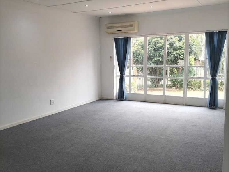 A spacious, empty living area featuring grey carpeting, recessed downlights, a wall-mounted air conditioning unit, and large glass sliding doors leading to the garden.