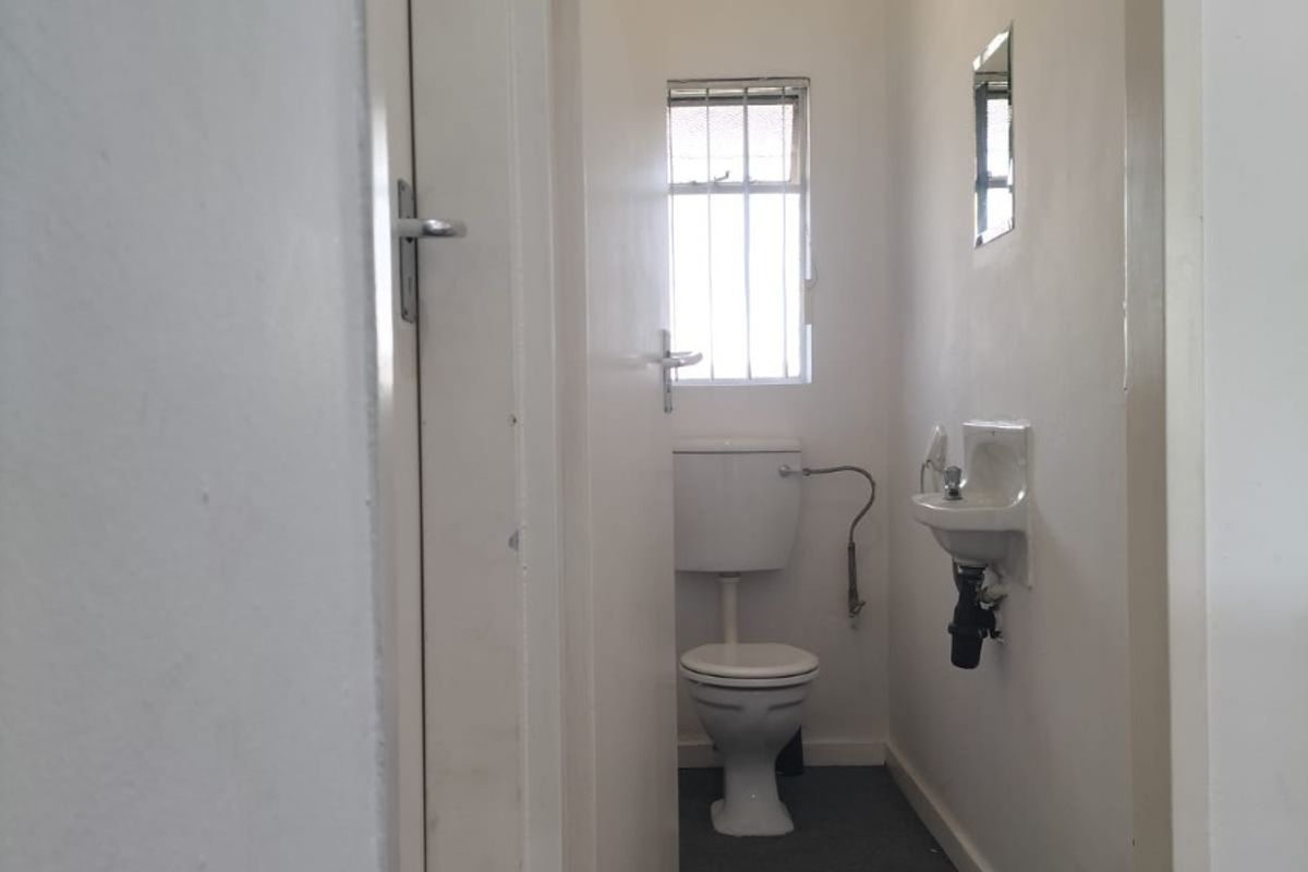 A compact, functional commercial restroom featuring a standard toilet, wall-mounted basin, and security-barred window, requiring cosmetic updates to improve its overall condition.