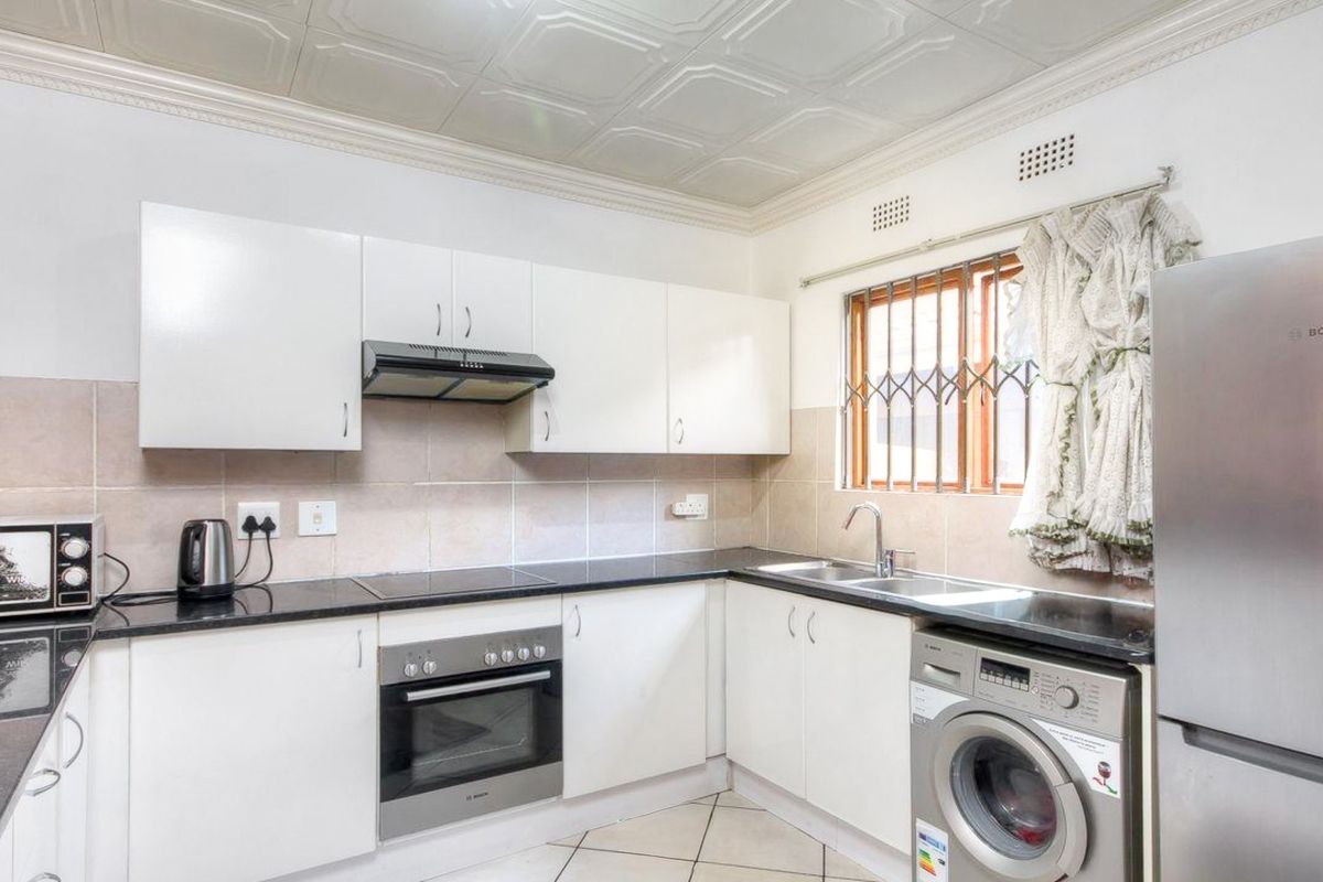 Functional kitchen featuring white cabinetry, granite countertops, built-in oven, hob, extractor fan, and tiled backsplash with a double sink and security window bars.