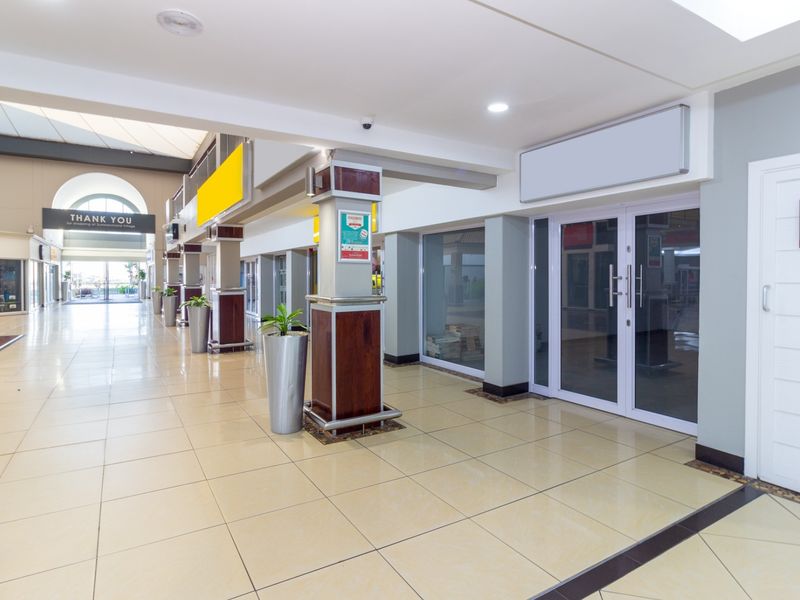 A clean, well-maintained retail shopping center corridor featuring tiled floors, structural columns, and glass-fronted commercial units.
