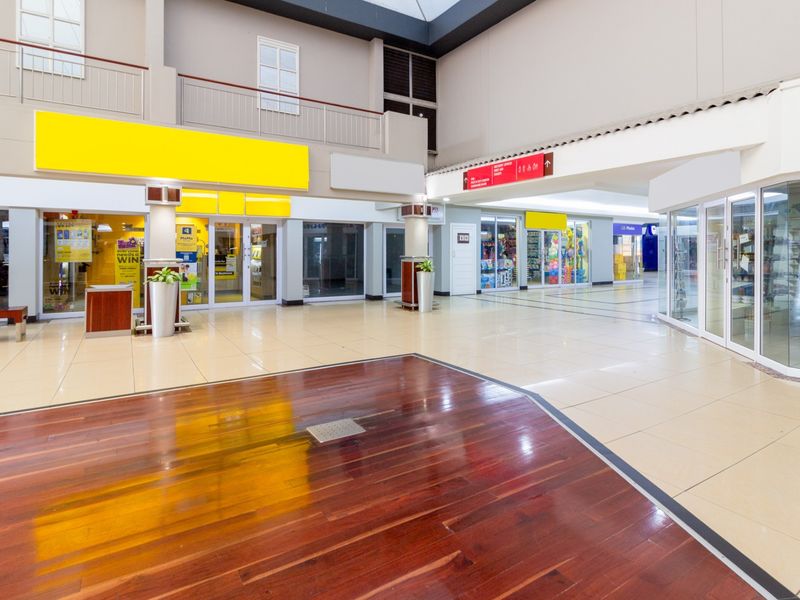 A bright, open-plan retail mall corridor featuring a mix of tiled and hardwood flooring, glass storefronts, and a mezzanine level with metal railings.