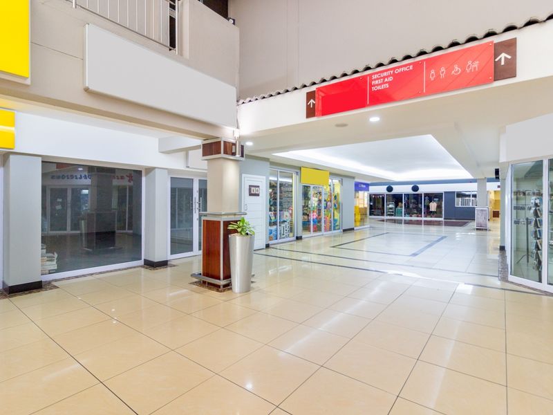 A well-lit retail shopping center corridor featuring tiled flooring, glass-fronted shop units, and clear directional signage for amenities.