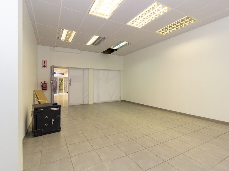 An empty open-plan retail space featuring ceramic floor tiles, a suspended ceiling with recessed lighting, and a fire safety installation.
