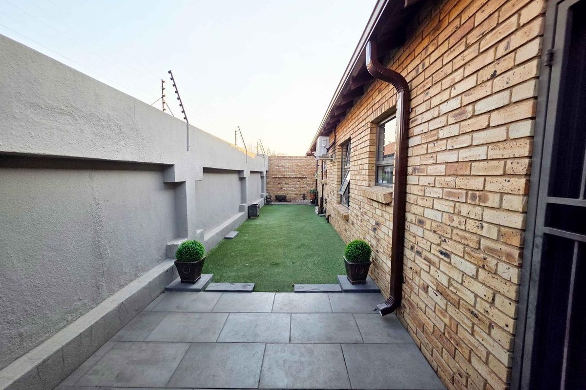 A neat rear exterior area featuring a tiled patio, artificial lawn, face brick facade, and secure boundary wall with electric fencing.
