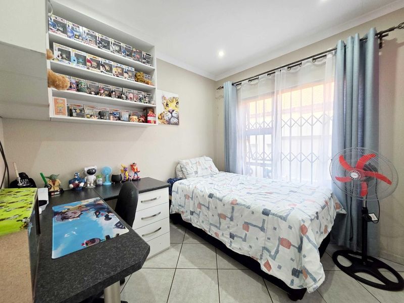 A functional bedroom featuring tiled flooring, built-in desk workspace, and wall-mounted shelving, with a large window fitted with a security gate.