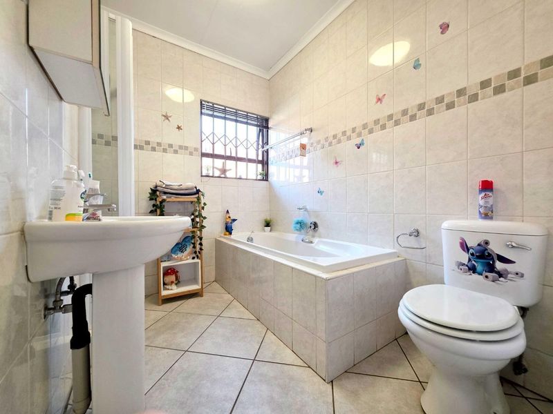 A functional bathroom featuring a pedestal basin, built-in bathtub, and toilet, finished with neutral wall and floor tiles and a security-fitted window.