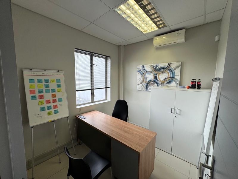 A functional office space featuring tiled flooring, a suspended ceiling with recessed lighting, and a wall-mounted air conditioning unit.
