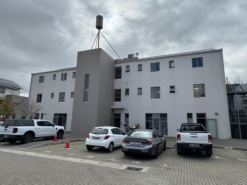 Front elevation of a multi-story office building featuring a paved parking area, external staircase, and rooftop telecommunications infrastructure.