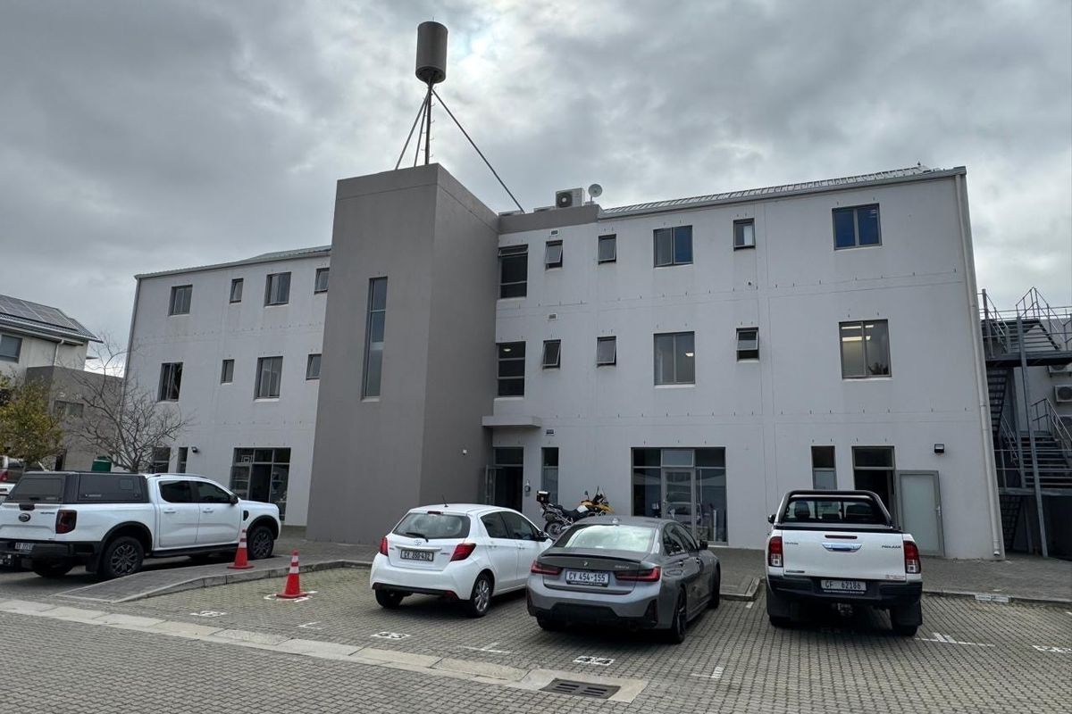 Front elevation of a multi-story office building featuring a paved parking area, external staircase, and rooftop telecommunications infrastructure.