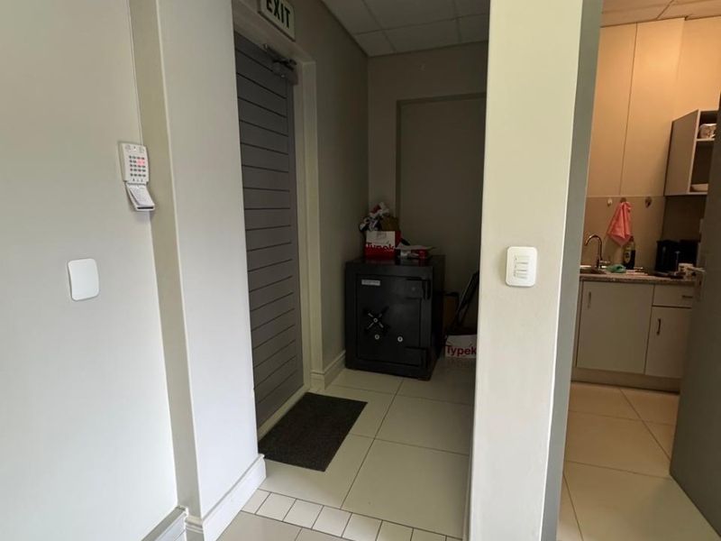 A functional office hallway featuring tiled flooring, a security alarm system, an exit door, and a view into an adjacent kitchenette area.