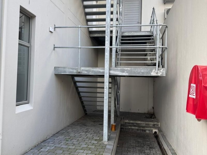 Rear exterior area featuring a galvanized steel staircase, paved walkway, drainage channel, and wall-mounted fire safety equipment.