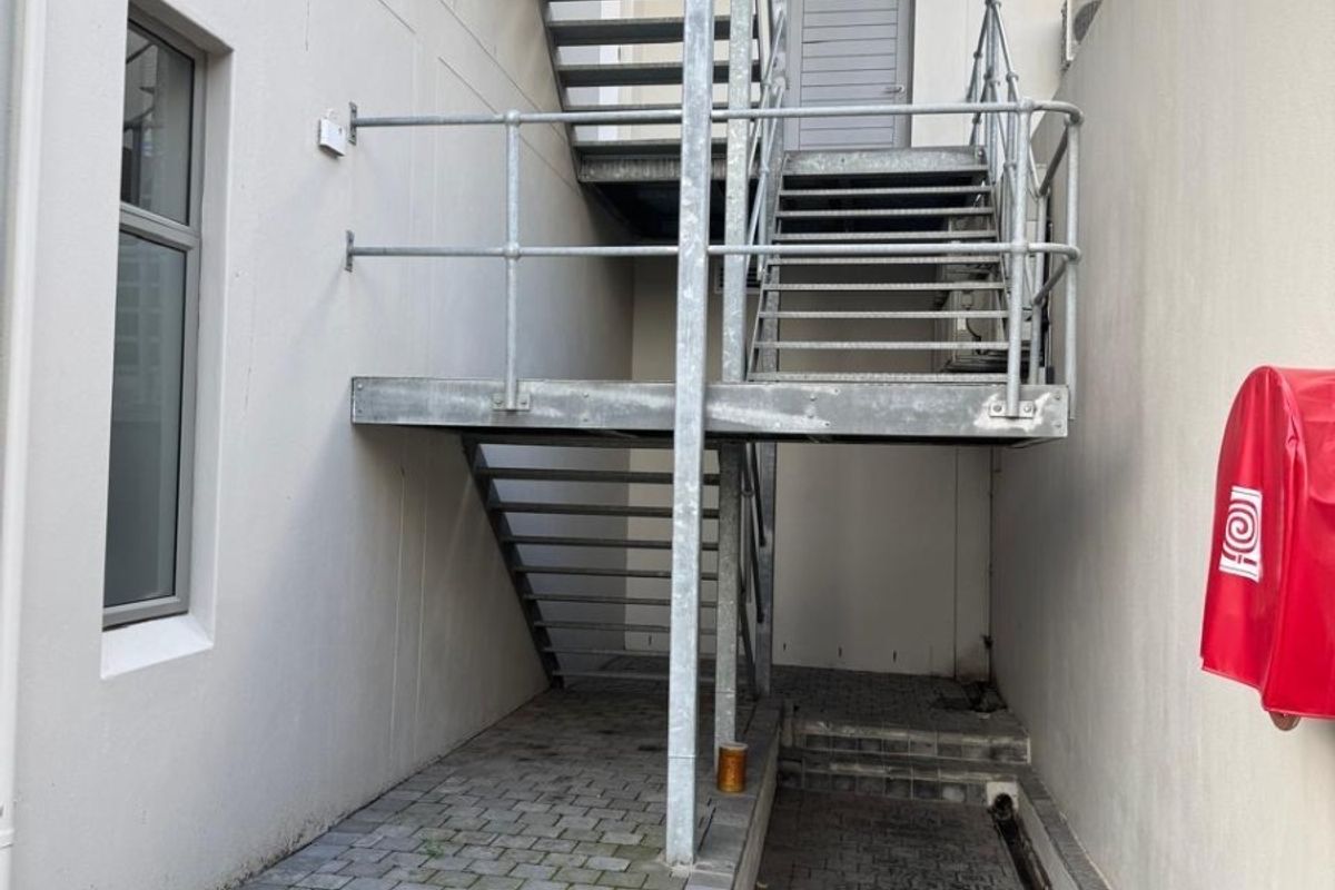 Rear exterior area featuring a galvanized steel staircase, paved walkway, drainage channel, and wall-mounted fire safety equipment.
