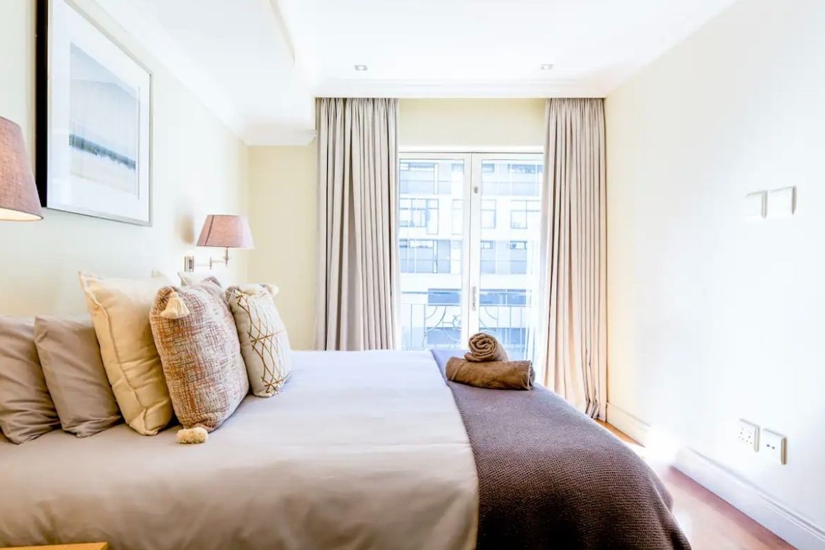 A bright and well-maintained bedroom featuring neutral tones, recessed ceiling lighting, and double glass doors leading to a balcony.