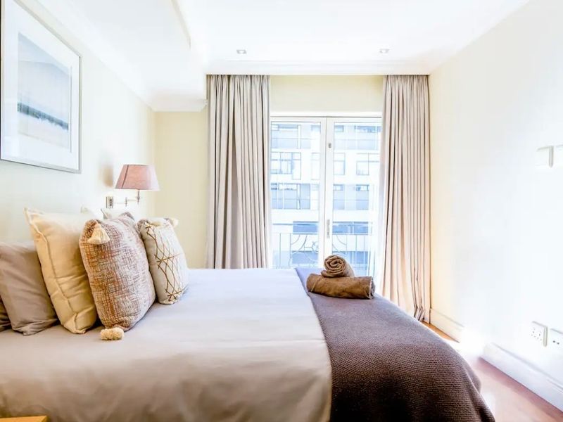 A bright and well-maintained bedroom featuring neutral tones, recessed ceiling lighting, and double glass doors leading to a balcony.
