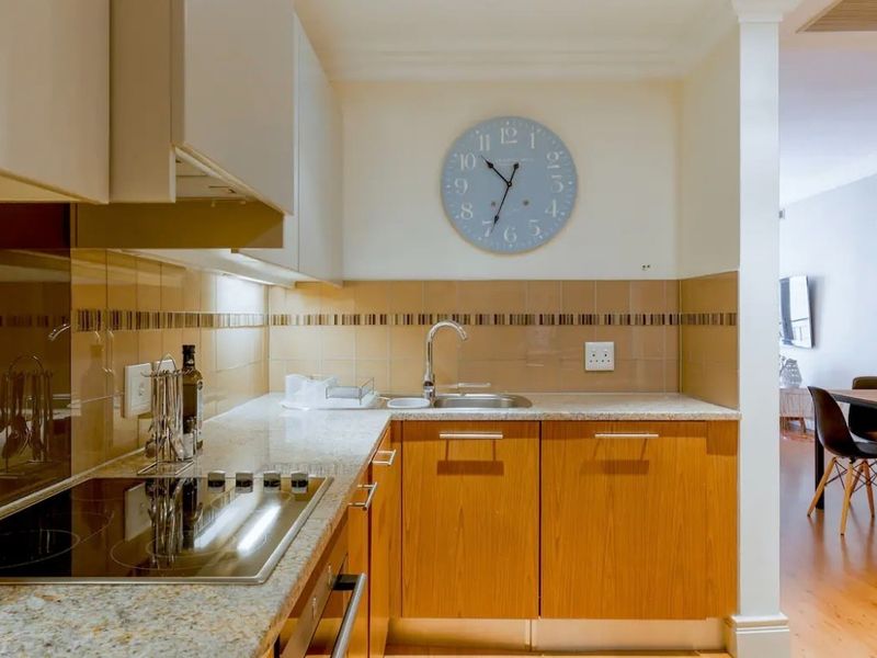 Compact kitchen featuring wood-finish cabinetry, granite countertops, tiled backsplash, and integrated electric hob and oven, flowing into the adjacent dining area.