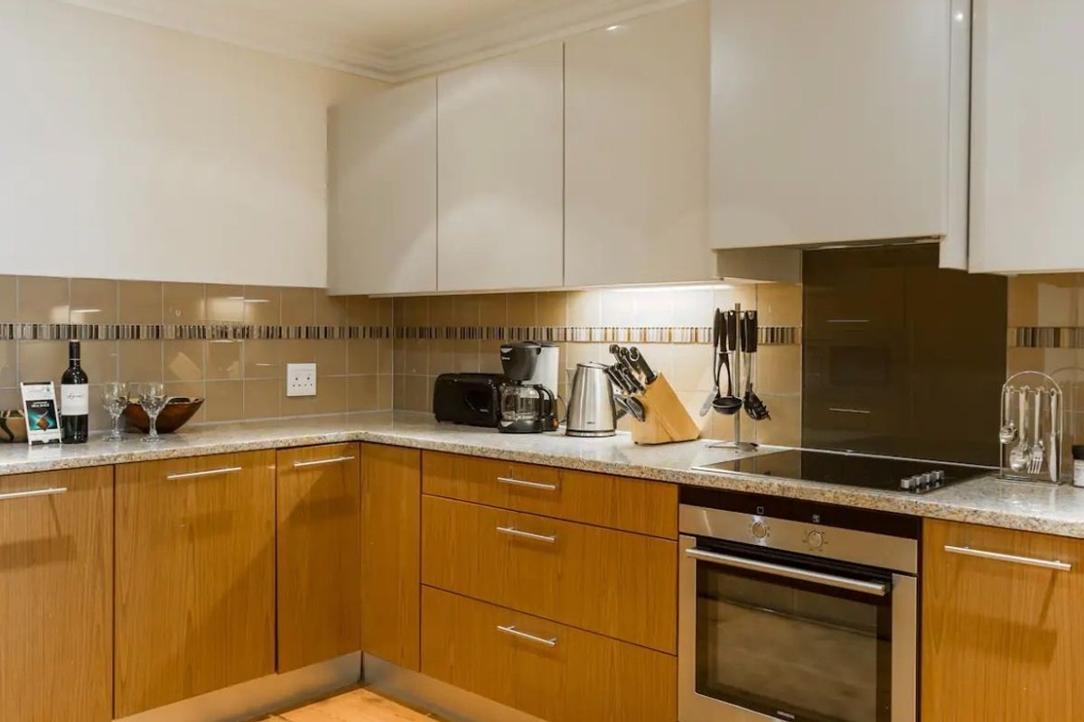 Modern kitchen featuring wood-finish lower cabinetry, neutral upper cabinets, granite countertops, and integrated stainless steel appliances with a tiled backsplash.