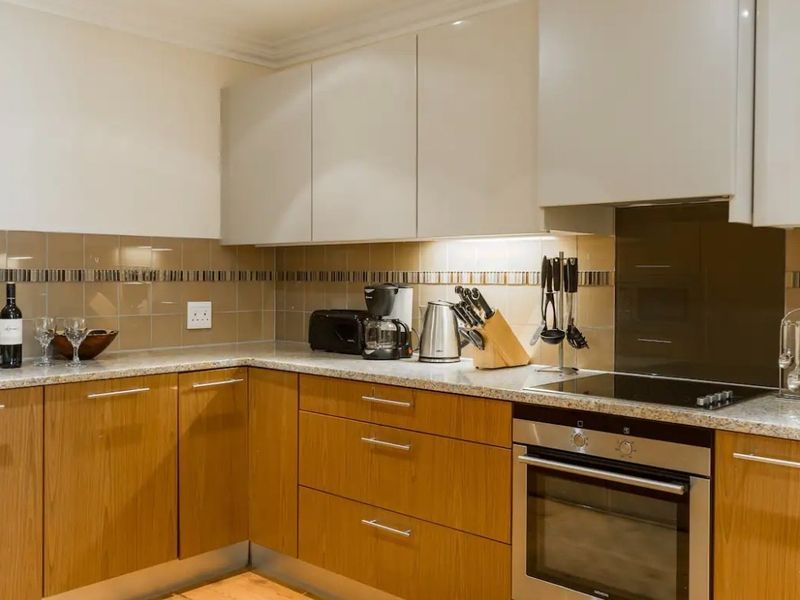 Modern kitchen featuring wood-finish lower cabinetry, neutral upper cabinets, granite countertops, and integrated stainless steel appliances with a tiled backsplash.