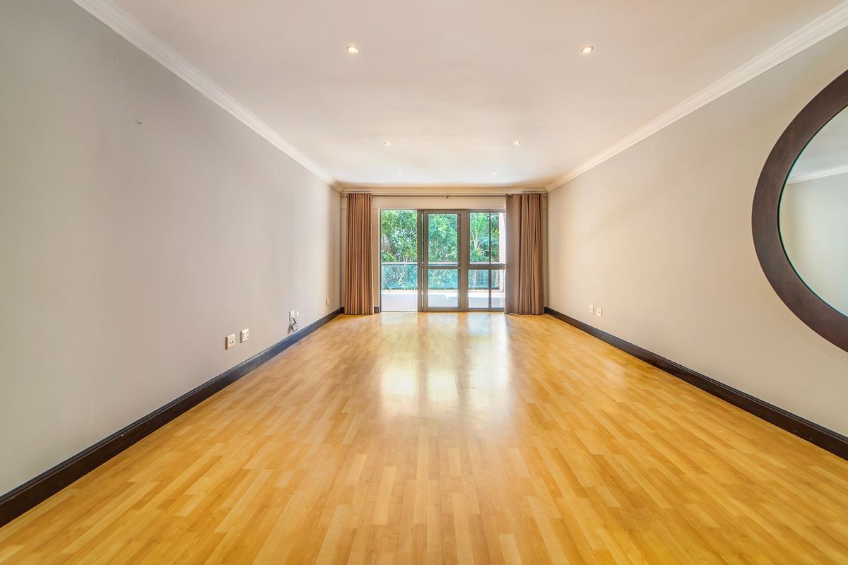 A spacious, empty living room featuring light laminate flooring, recessed downlighting, and a large sliding glass door providing access to the balcony.