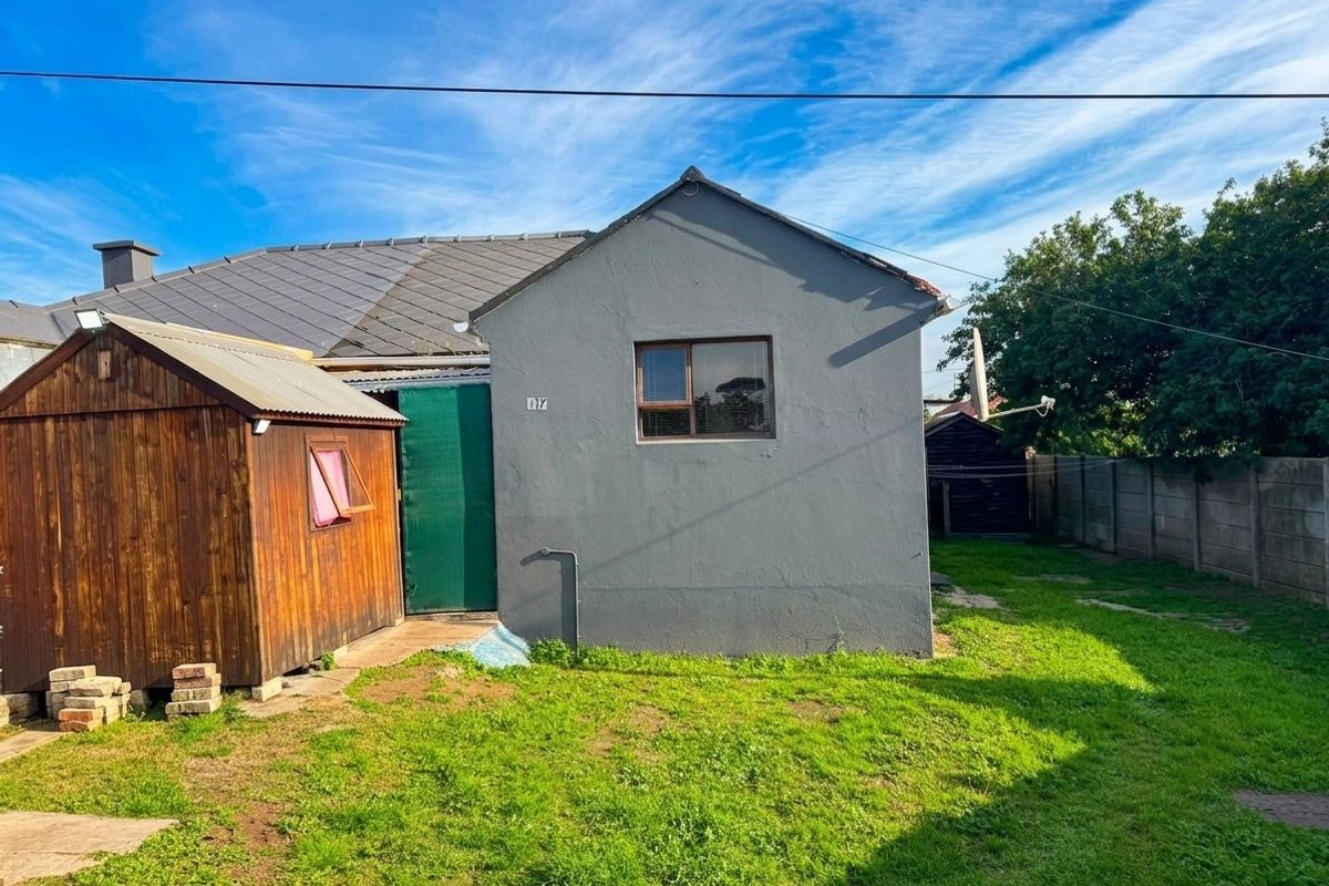 Rear view of the property featuring a grey rendered exterior, a timber garden shed, and a grassed backyard area with concrete boundary walling.