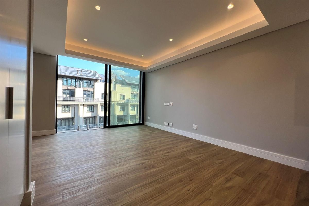 A bright, modern, and empty living room featuring recessed cove lighting, wood-look laminate flooring, and large sliding glass doors leading to an exterior balcony.
