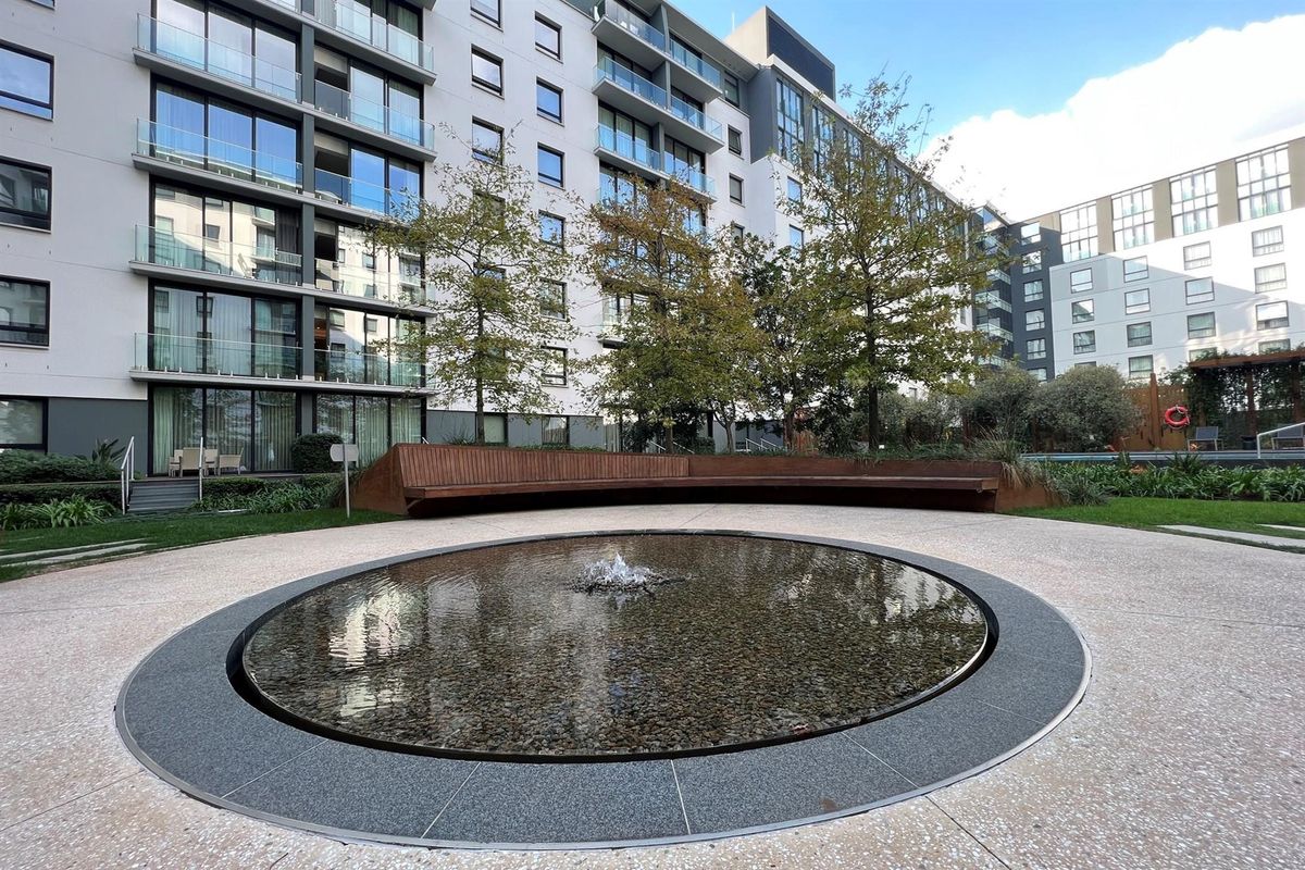 A well-maintained communal courtyard featuring a circular water feature, curved timber seating, and landscaped gardens set against a modern multi-story residential building.