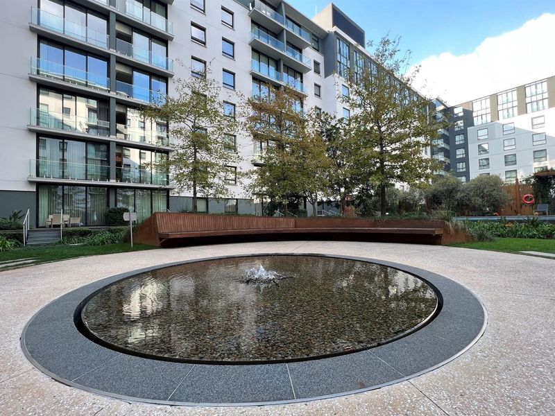 A well-maintained communal courtyard featuring a circular water feature, curved timber seating, and landscaped gardens set against a modern multi-story residential building.