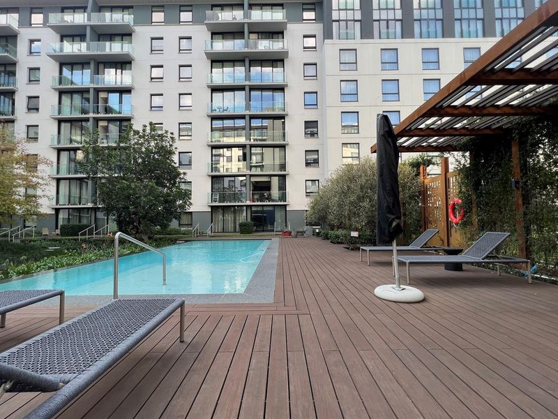 A well-maintained communal swimming pool area featuring timber decking, a pergola, and landscaped surroundings set against a modern apartment building.