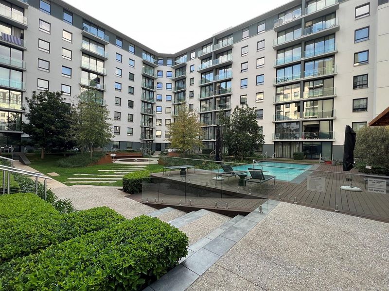 A well-maintained communal pool area featuring timber decking, glass balustrades, and landscaped gardens set within a modern multi-story residential complex.