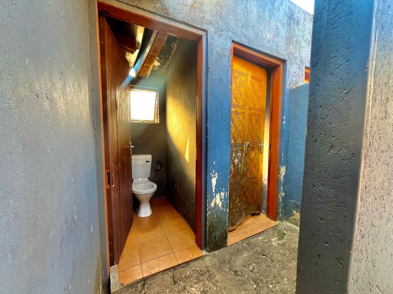 Exterior view of a small outdoor bathroom facility featuring a toilet, tiled floor, and timber doors, showing signs of significant wear and weathering.