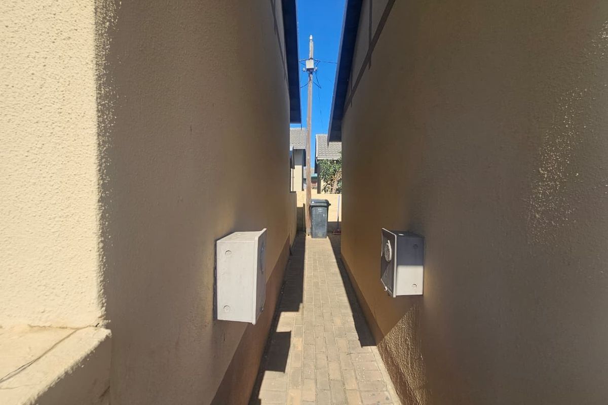A narrow paved side passage between two townhouse structures featuring exterior electrical meter boxes mounted on the plastered walls.