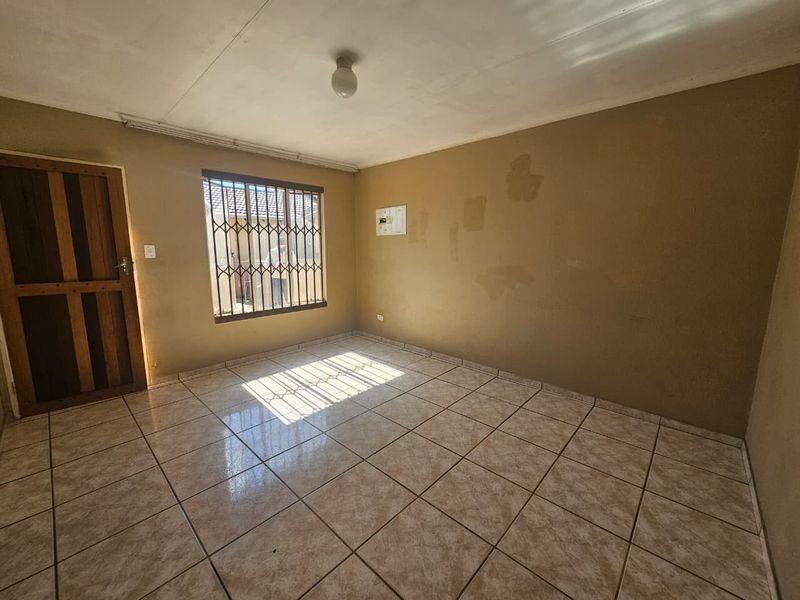 An empty living room featuring tiled flooring, security bars on the window, and a wooden door, requiring cosmetic updates to the wall finishes.