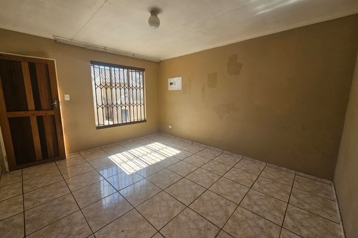 An empty living room featuring tiled flooring, security bars on the window, and a wooden door, requiring cosmetic updates to the wall finishes.