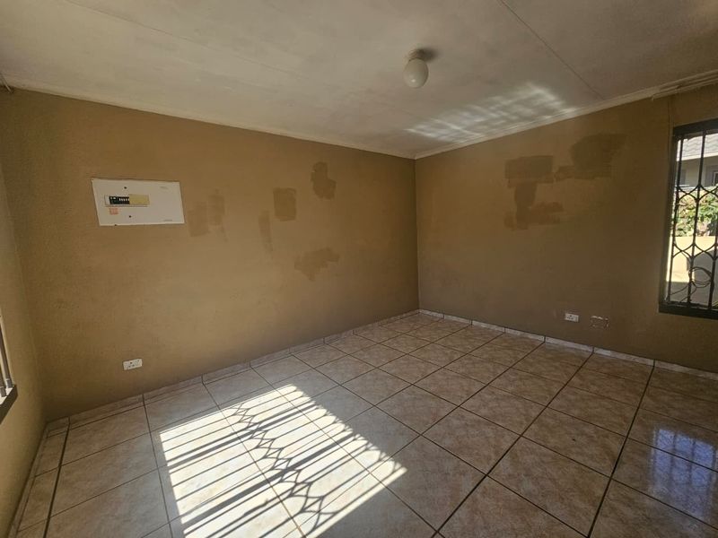 An empty living room featuring ceramic floor tiles, security bars on windows, and an integrated electrical distribution board, requiring cosmetic wall repairs.