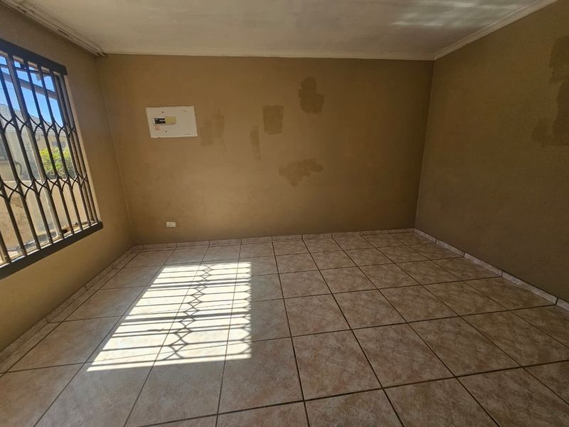 An empty living room featuring tiled flooring, security bars on the window, and an electrical distribution board, requiring cosmetic wall updates.