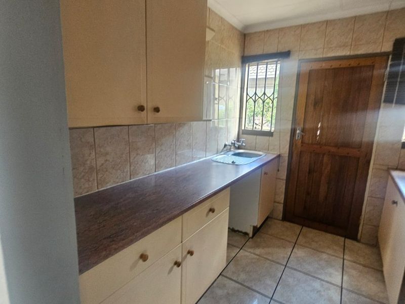 A functional kitchen featuring tiled flooring, built-in cabinetry with laminate countertops, a stainless steel sink, and direct access to an exterior timber door.