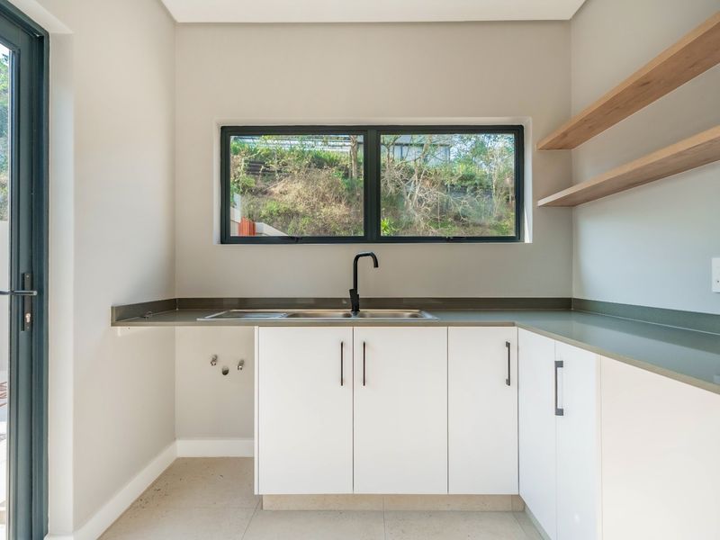 A clean and modern scullery featuring white built-in cabinetry, stone countertops, a stainless steel sink, and floating timber shelving with direct outdoor access.
