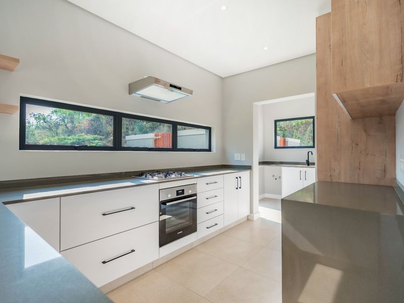 Modern kitchen featuring sleek white cabinetry, stone countertops, built-in oven, gas hob, and an adjoining scullery area with neutral tiled flooring.