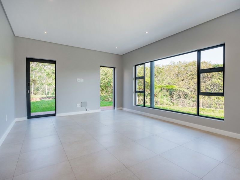 A bright, modern, and spacious living area featuring large format floor tiles, recessed lighting, and expansive windows with views of the surrounding greenery.